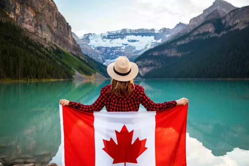 Mujer Vestida Con Camisa De Vestir A Cuadros Roja Y Negra Y Sombrero Fedora Beige Sosteniendo La Bandera De Canadá Mirando Al Lago