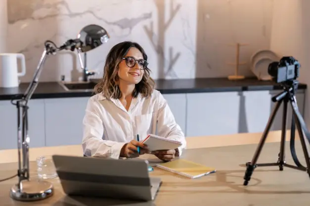 Joven mujer con gafas trabajando en interiores con portátil y cámara, sonriendo mientras toma notas. (Foto: Pexels)