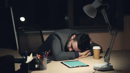 Hombre con traje de oficina durmiendo en el escritorio con café y tablet cerca, simbolizando la fatiga laboral. (Foto: Pexels)