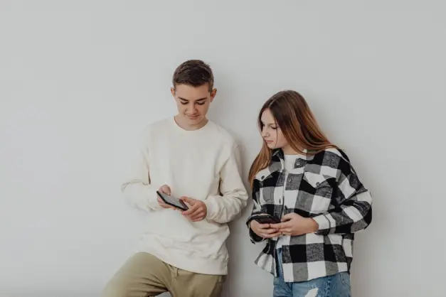 Chico y chica adolescentes usando smartphones mientras se apoyan en una pared blanca en interiores. (Foto: Pexels)