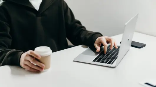 Configuración informal de oficina en casa con una persona escribiendo en una laptop mientras sostiene una taza de café. (Foto: Pexels)