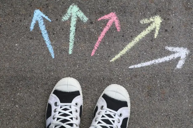 Zapatillas negras sobre el pavimento con flechas de tiza coloridas apuntando en diferentes direcciones. (Foto: Pexels)