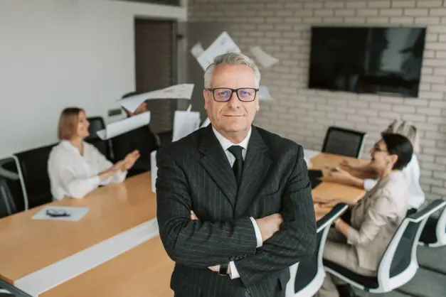 Ejecutivo de negocios de pie con confianza en la sala de reuniones, con el equipo involucrado en una discusión detrás. (Foto: Pexels)