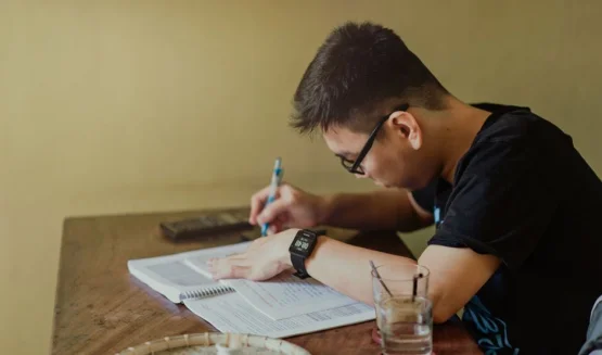 Joven sentado frente a un escritorio mientras estudia y toma notas en una hoja.