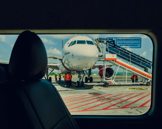 Avión visto desde un coche, estacionado en la pista de un aeropuerto con la tripulación trabajando cerca.