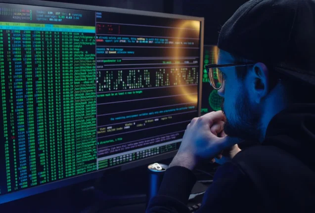 Hombre trabajando intensamente en programación de computadoras con código mostrado en monitores duales en una habitación poco iluminada.