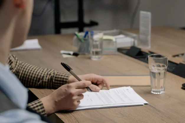 Persona con atuendo de negocios firmando un documento en una mesa de madera en un entorno de oficina.