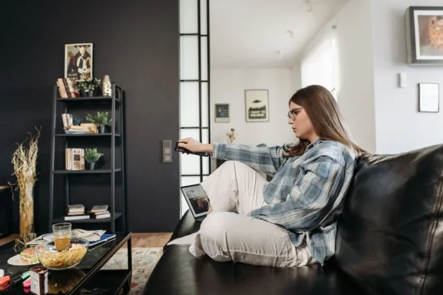 Mujer sentada en un sofá en una sala de estar acogedora, sosteniendo un control remoto, rodeada de aperitivos y decoración.
