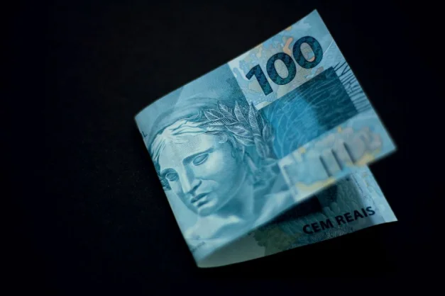 Un billete de 100 reales brasileños mostrado contra un fondo negro, destacando su diseño y color.
