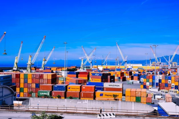 Un bullicioso puerto de carga con coloridos contenedores apilados y numerosas grúas bajo un cielo azul claro.