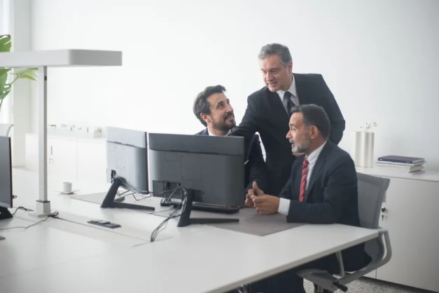 Tres ejecutivos de negocios con trajes colaborando alrededor de monitores dobles en un entorno de oficina moderna.