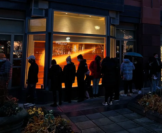Individuos esperando en la fila fuera de una tienda bien iluminada por la noche, posiblemente durante un evento de ventas.