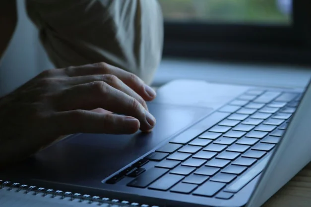 Primer plano de las manos de una persona escribiendo en el teclado de una computadora portátil en un interior con iluminación tenue.