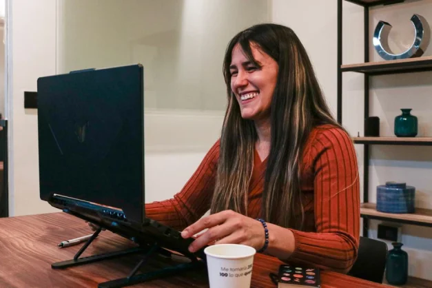 Mujer sonriente con suéter rojo disfrutando de una videollamada en su oficina en casa, equipada con computadora portátil y café.