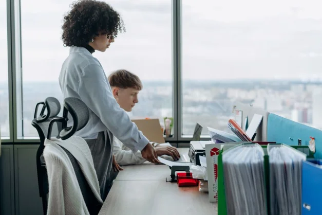 Dos compañeros colaboran en una oficina moderna con vista a la ciudad, mejorando el trabajo en equipo y la productividad.